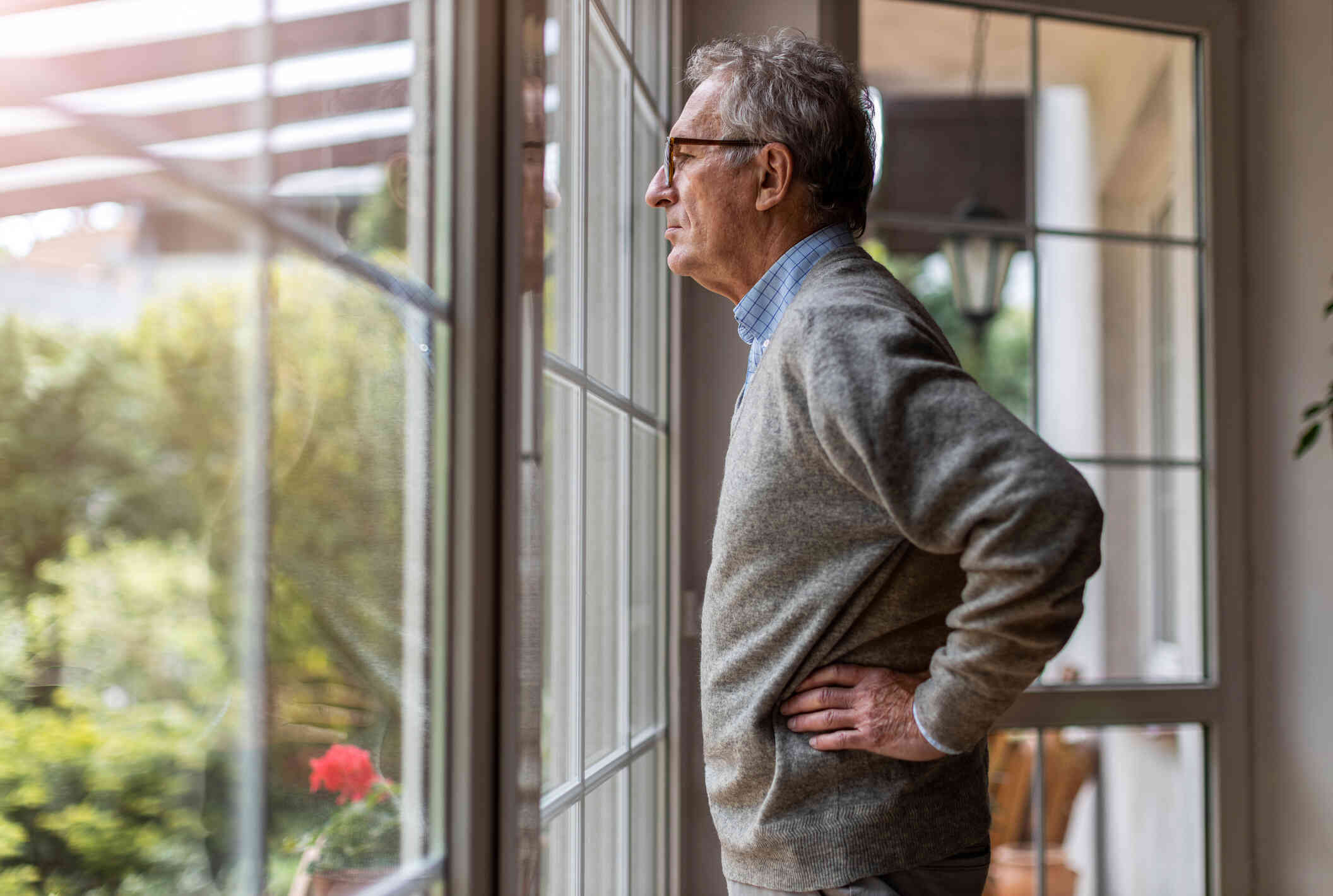 An elderly man in a grey sweater stands near the window of his home and rests his hands on his hip while deep in thought.
